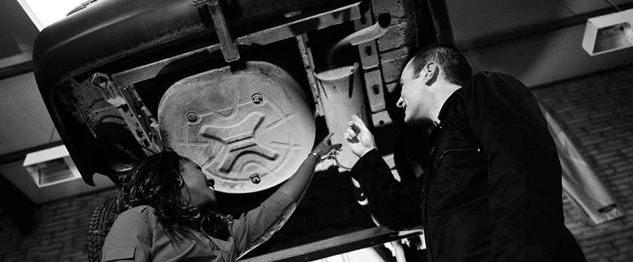 An image of two people in a mechanic's shop pinting to the underside of a hoisted car