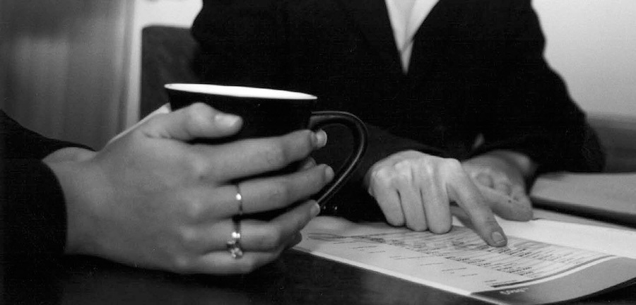 A photo of one person holding a cup of coffee as another person explains a document to them.